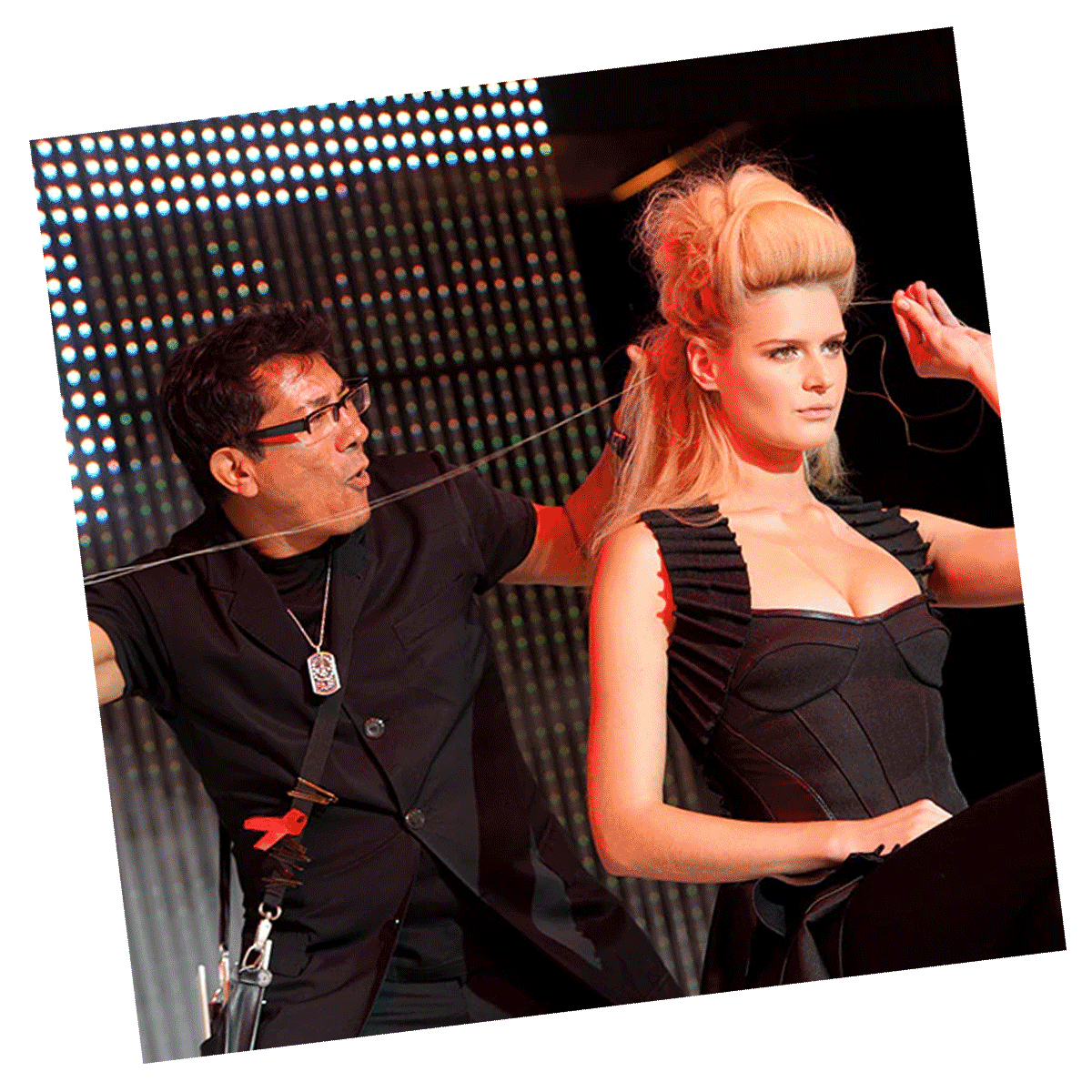 Sam Villa styling a model's voluminous updo hairstyle on stage during a live demonstration. The model is wearing a black outfit, and the background features bright stage lights.
